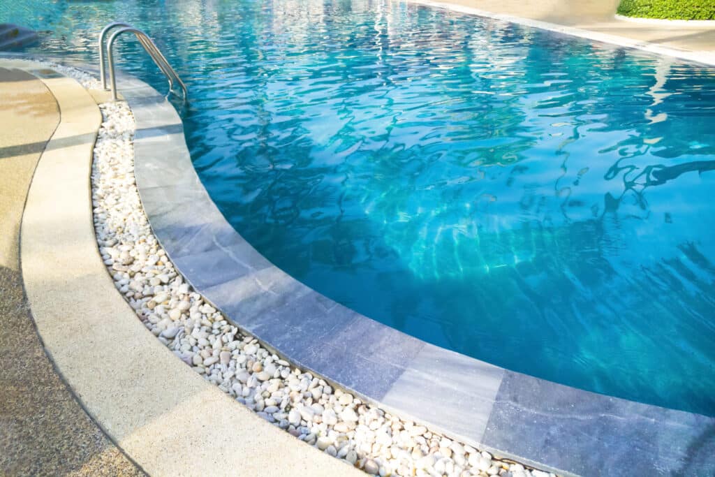 Piscine bleue étincelante : relaxation au bord de l'eau Piscine bleue incurvée avec main courante, bordée de galets blancs et d'une terrasse beige. L'eau claire reflète la lumière du soleil.