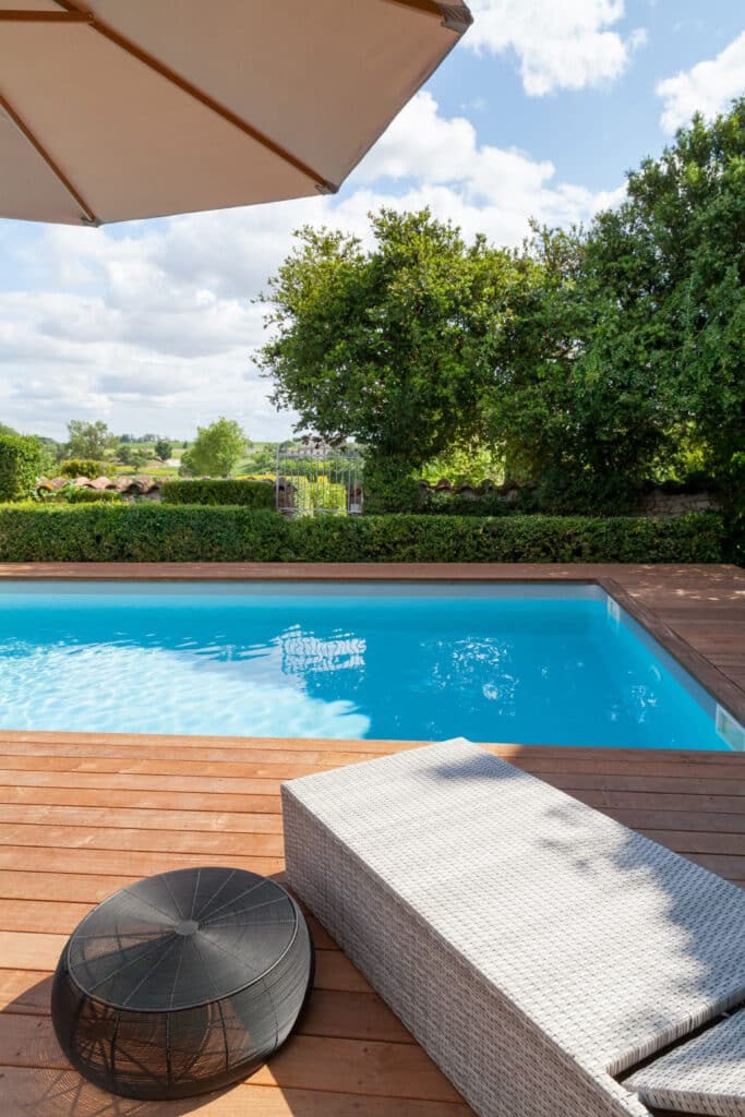 Piscine avec terrasse bois: détente dans jardin luxuriant Piscine moderne avec terrasse en bois, transat et table d'extérieur sous parasol. Jardin luxuriant et campagne sous ciel bleu.